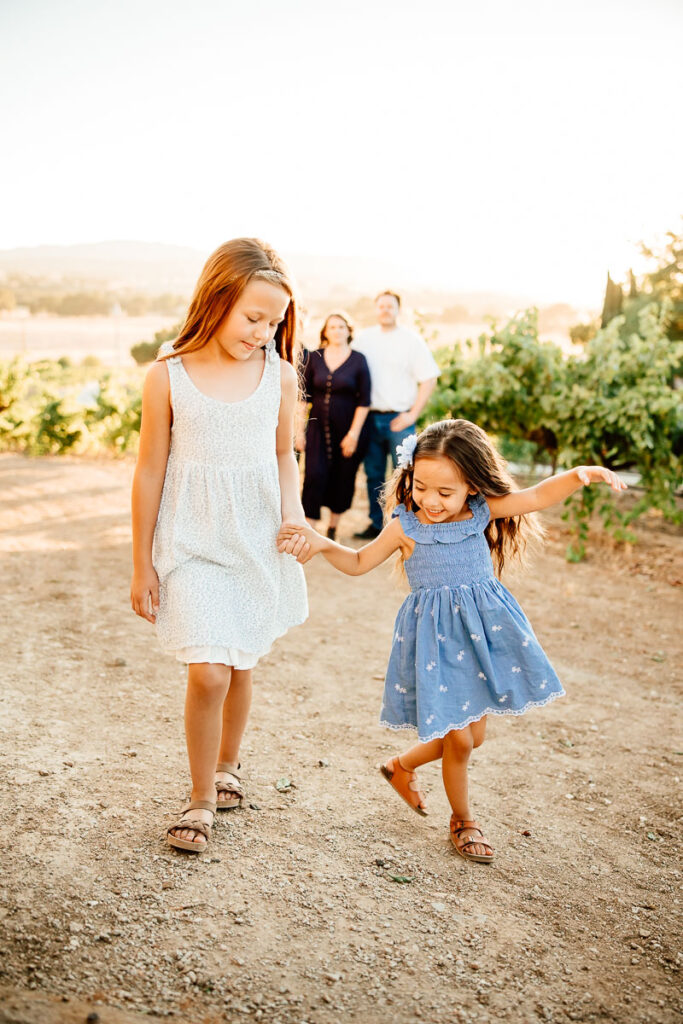 2 young girls dancing in a vineyard while their family looks on for a blog post of the importance of hiring a family photographer