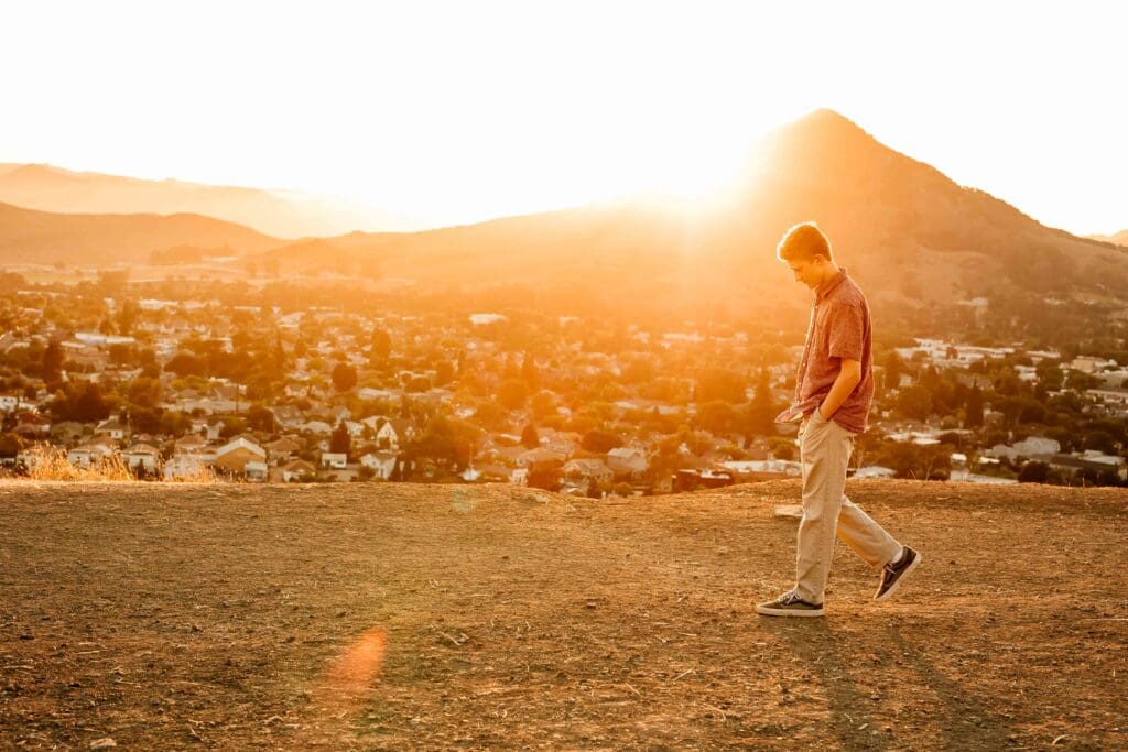 backlit image of a senior boy walking on a hill with his hometown behind him