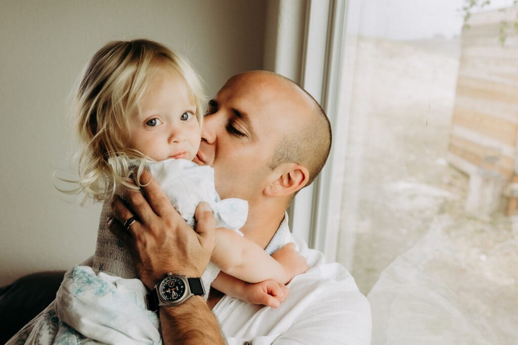Dad holds young daughter and kisses her on the cheek in a photo for a blog about in home lifestyle photo session. 